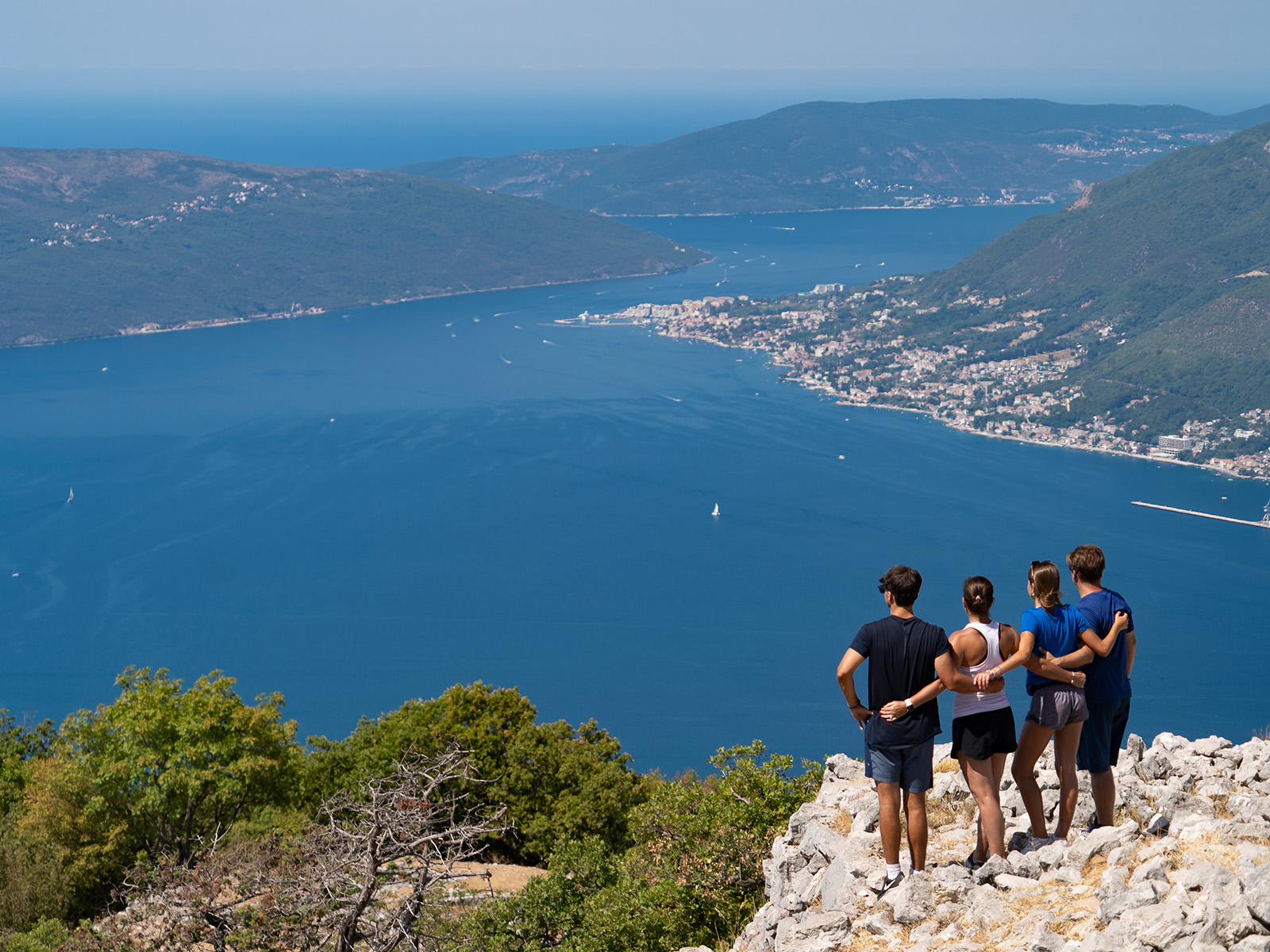 Four guests, arms around each other on hilltop, overlooking ocean vista.