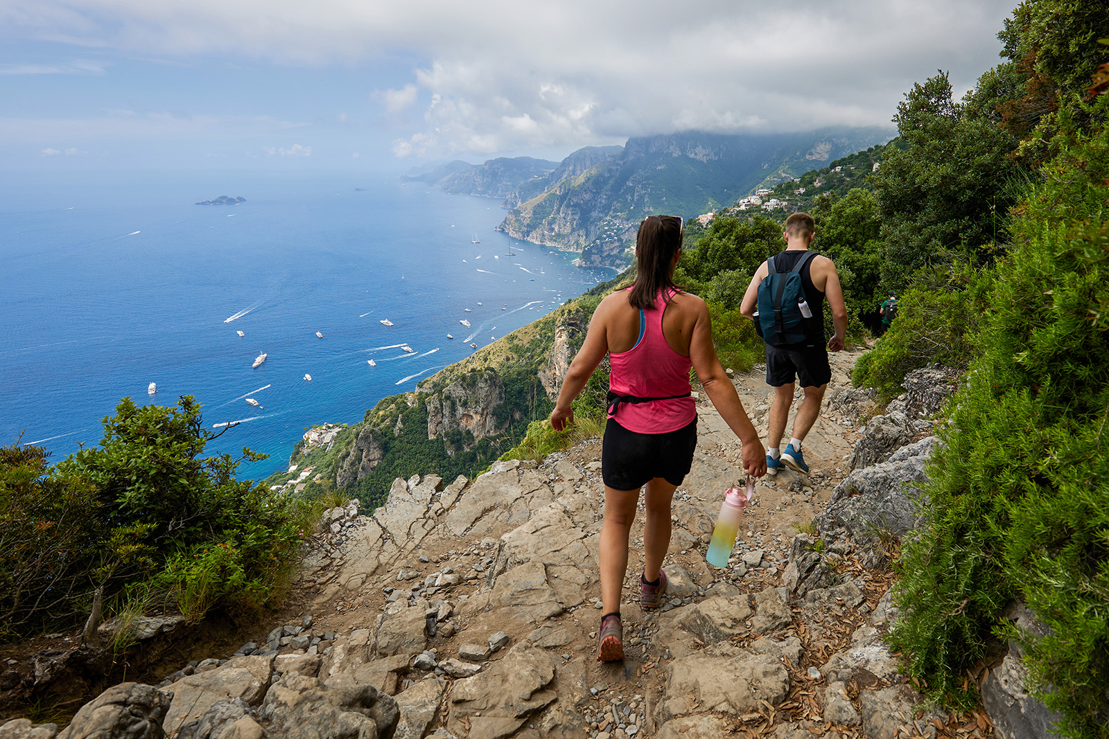 Two guests hiking down cliffside, boats in ocean down below.