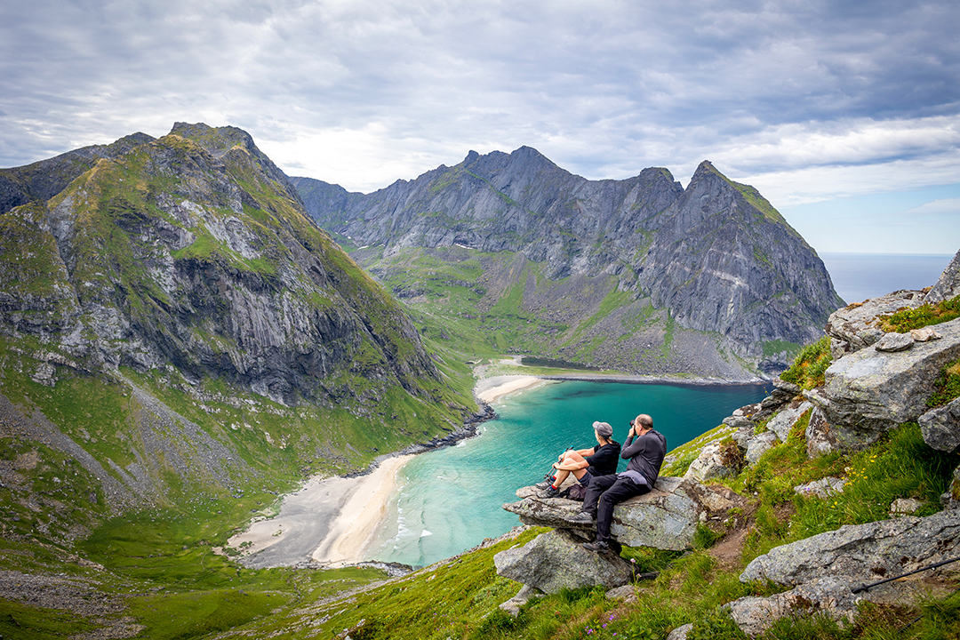 Man and woman on a rocky cliff, looking out to towards larger mountains an a beach