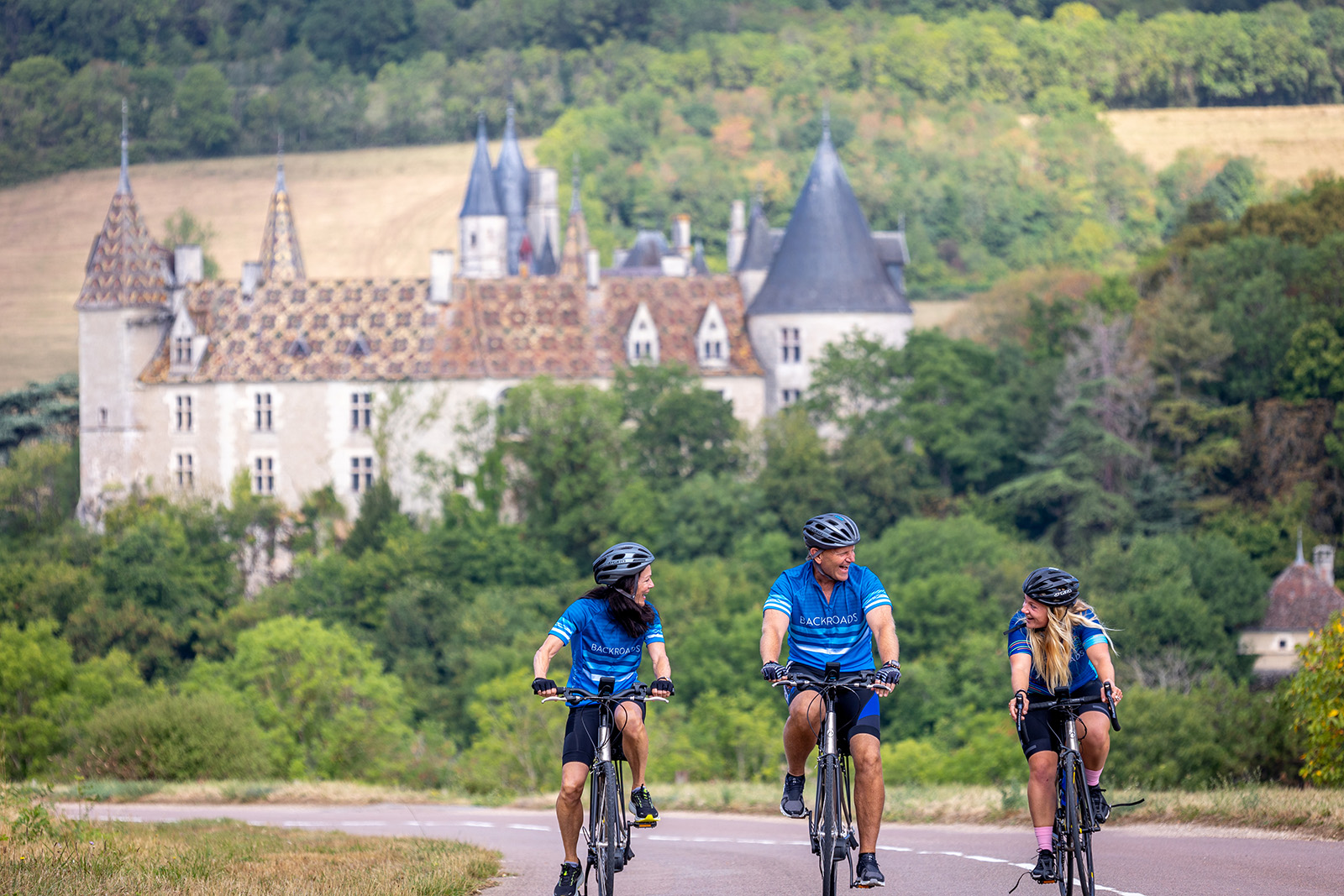 Two women and one man biking, with stone buildings and trees in the background