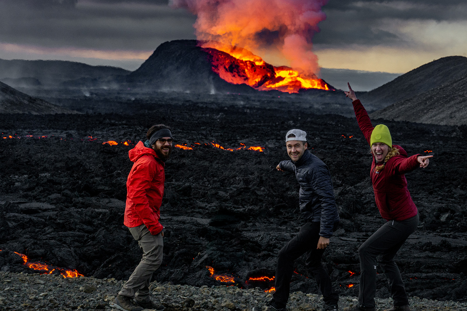 Three people smiling and pointing at an active volcano