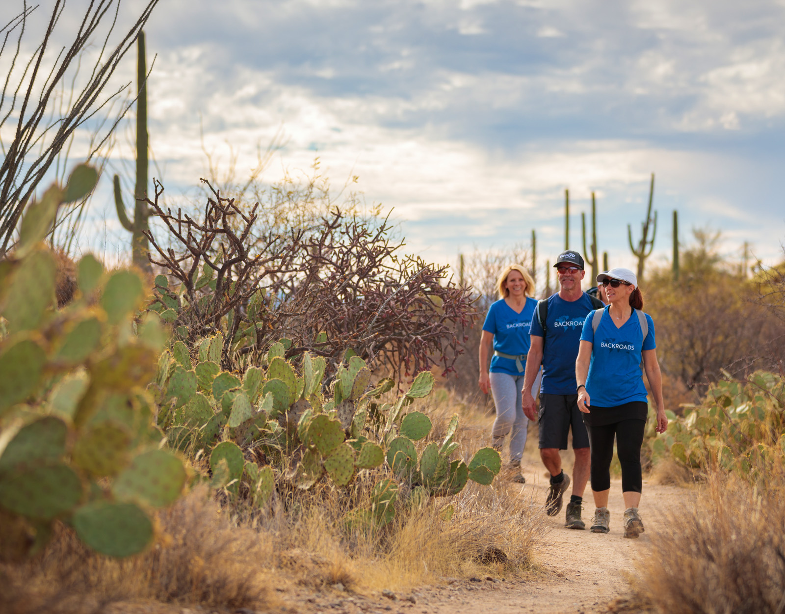 Two women and one man hiking on a dirt trail surrounded by cacti and dried trees