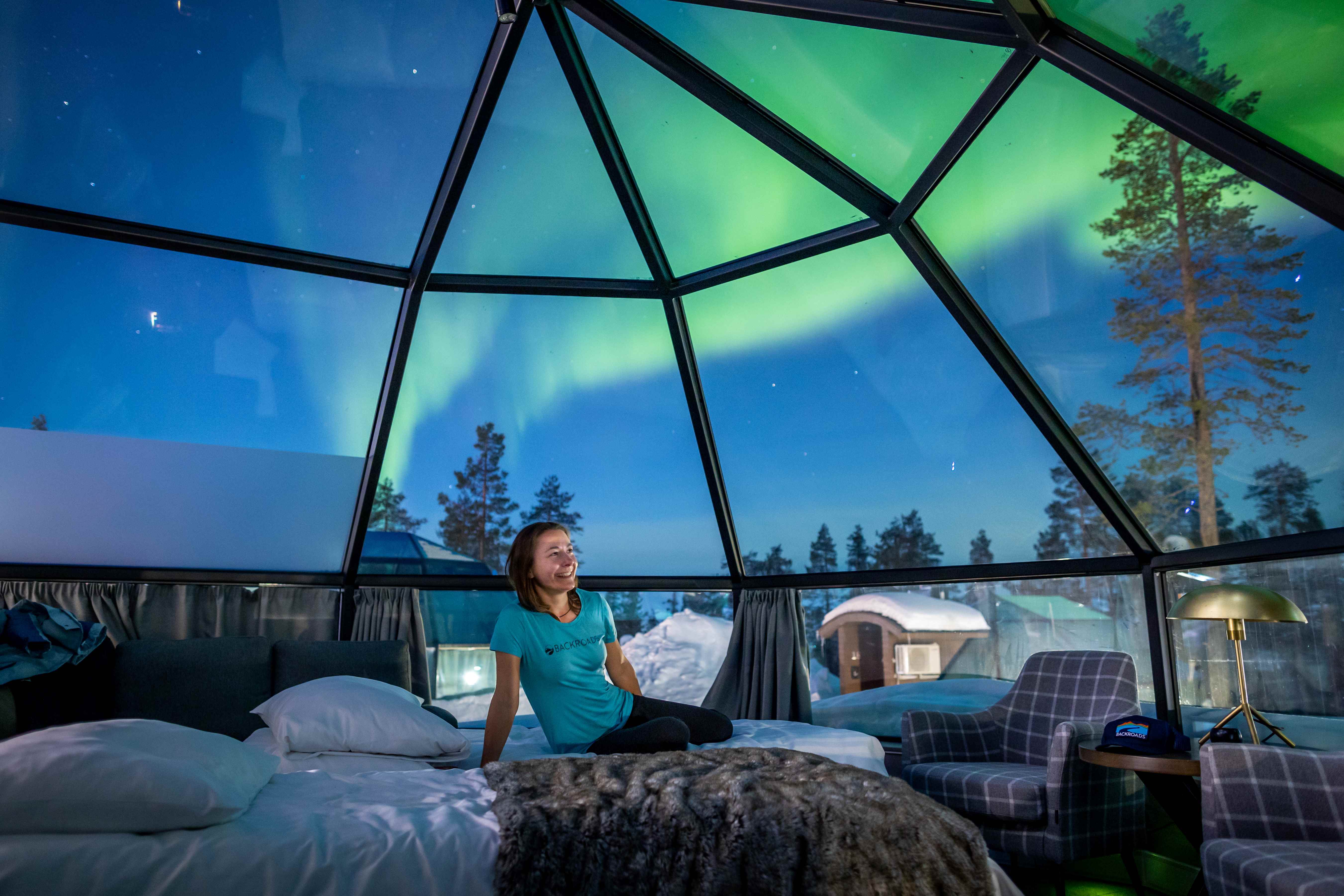 Woman sitting on a bed in a glass dome, with the Northern Lights in the sky