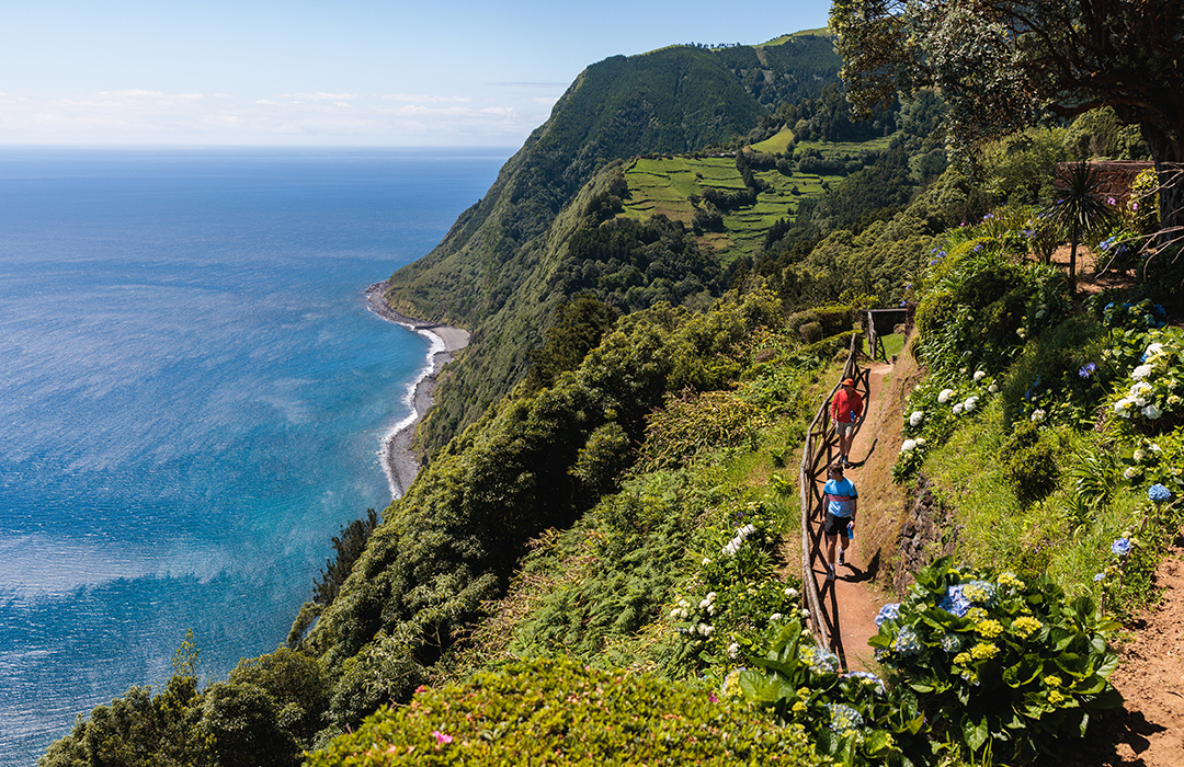 Two men walking along a dirt path on a cliff, with the ocean coast on the ground floor