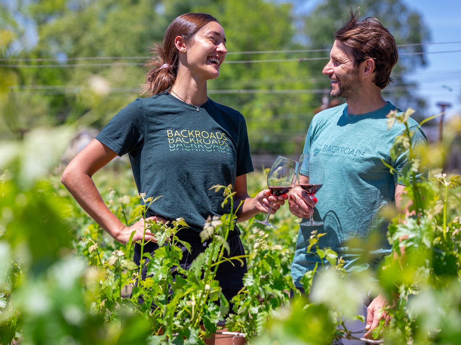Man and woman smiling while holding up wine glasses, standing in a vineyard