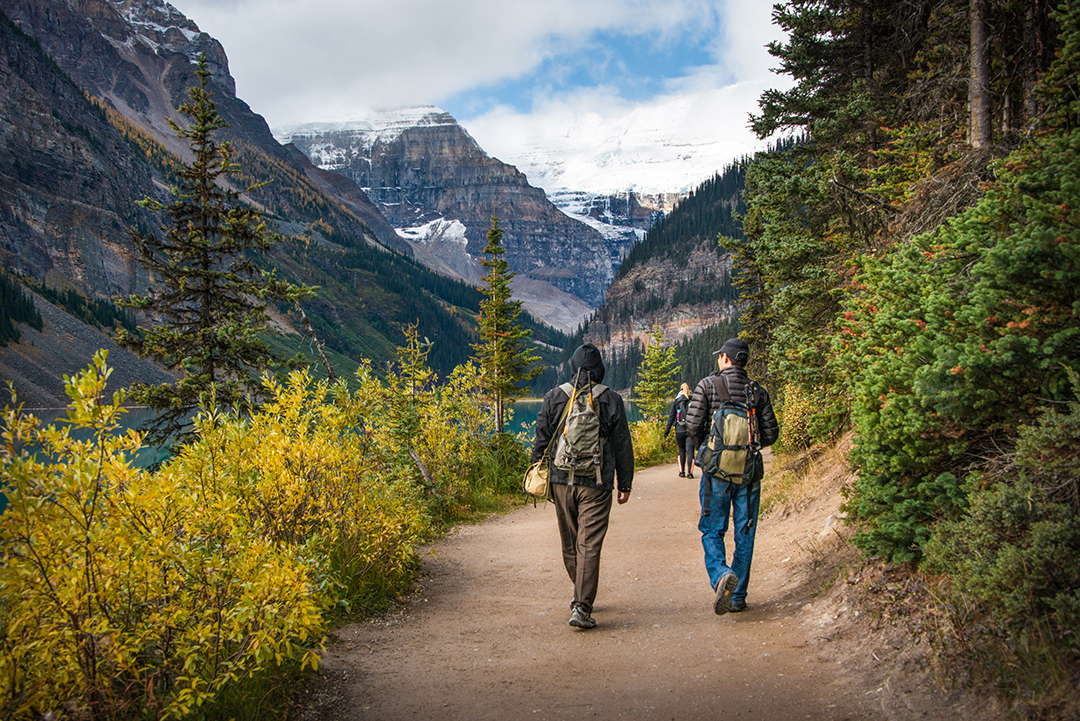 Two people hiking on a dirt trail, surrounded by bushes and mountains in the distance