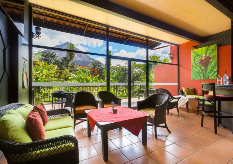 Bar and lounge with woven chairs and large glass walls and doors leading to a jungle