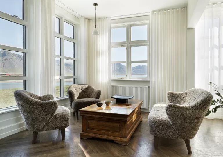 Corner of a white room with fuzzy, cushioned chairs and large windows looking out to tall mountains