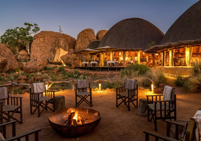 Woven chairs surrounding a fire pit, with straw huts in the background