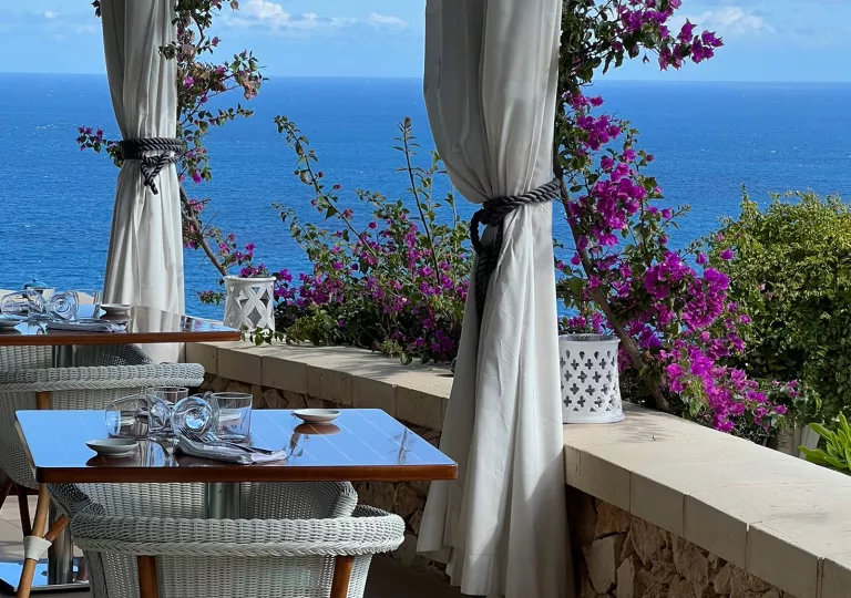 Outdoor dining area with wooden tables and woven white chairs, with the ocean in the background