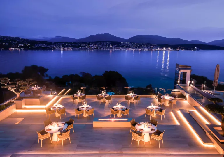 Illuminated outdoor patio with woven chairs and white, circular tables, looking out towards a large lake
