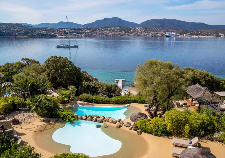Two outdoor pools surrounded by tall trees, with views of a lake and distant town in front