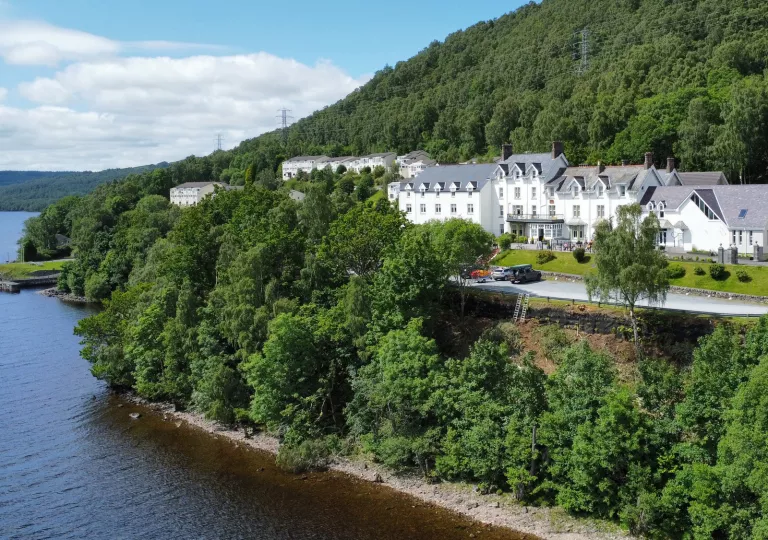 Exterior view of white hotel complex, surrounded by tall trees with a lake on the ground level