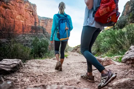 Two guests walking down desert trail, orange cliffs, bushes beside them.