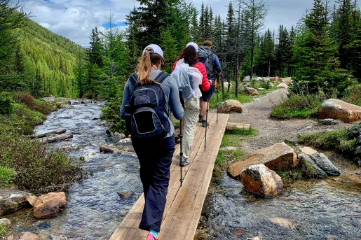 Three guests hiking over stream, forest, clouds, hillside in distance.