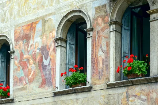Shot of ancient mural on side of building, open windows, flowers.