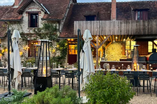 Outdoor dining area in front of a tan and brown brick building, with portable space heaters and umbrellas in the background