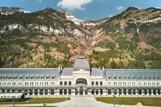 Wide tan and gray building with a large, gray courtyard and tall mountains in the background