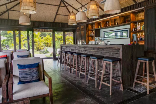 Indoor bar with a wooden counter and stools, with cushioned chairs in the background