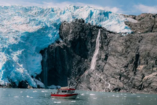 Red and gray boat floating next to large glaciers and cliffs