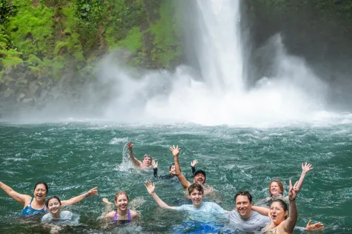 Large family smiling and laughing while swimming under a waterfall in a pond