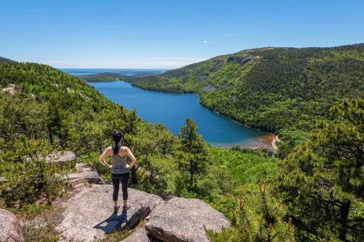 Woman standing on top of a boulder, looking out to a large lake and forest