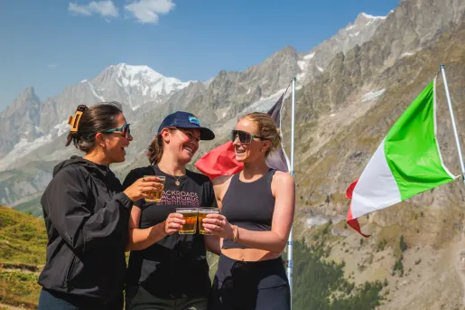 Three women smiling next to an Italian flag, with large mountains in the background