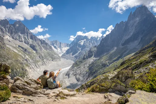 Man and woman sitting on a cliff, pointing out to large mountains in the distance