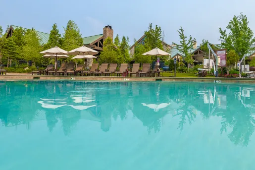 Outdoor pool surrounded by beige and brown chairs and umbrellas, with buildings covered by trees