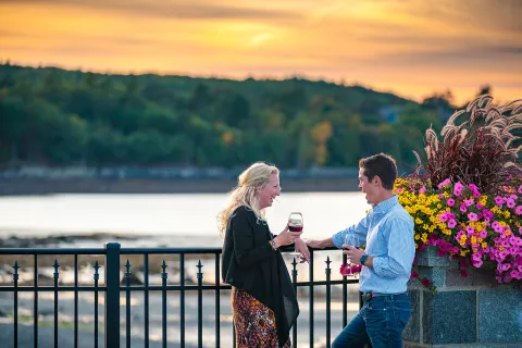 Two guests chatting, wine glasses in hand, sunset and beach in background.