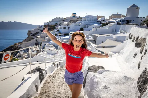 Guest emoting to camera, white walled coastal buildings behind her.