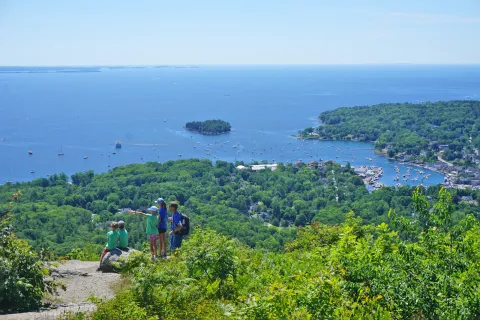 Wide shot of a coastal vista, sailboats visible in water, group of young guests in foreground.