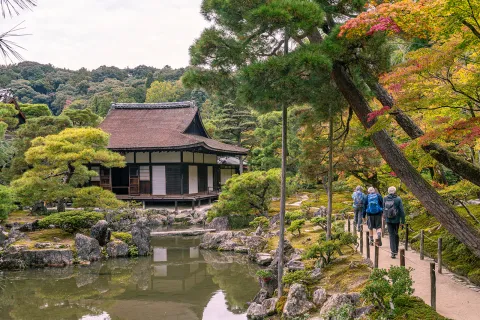 Three people walking on a small path on the right towards a Japanese-style building in the forest