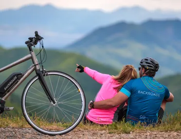 Two guests on roadside, pointing towards horizon, bike behind them.