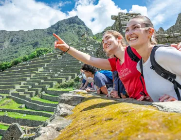 Two women smiling and pointing towards the sky, with large stone ruins in the background